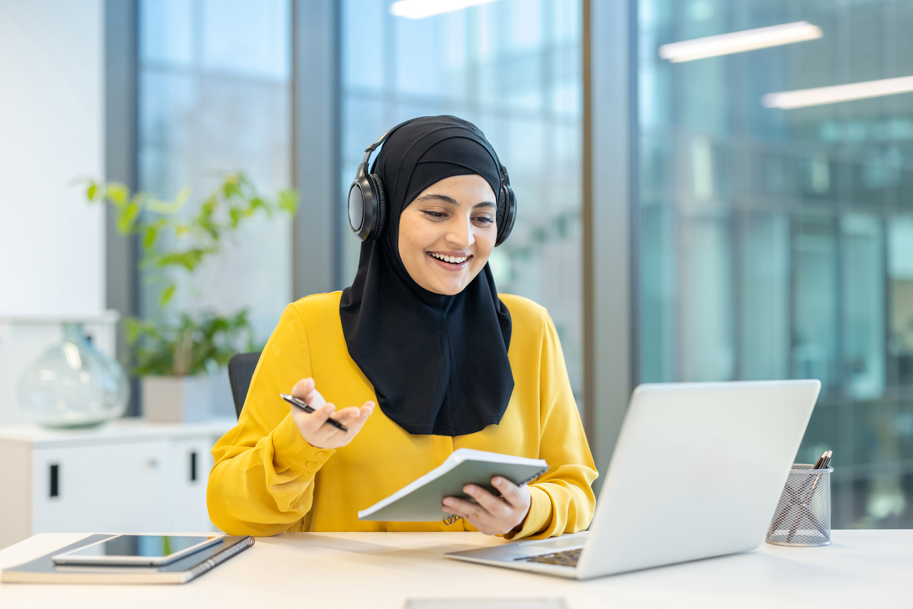 © Liubomir | stock.adobe.com | A smiling woman in a hijab, wearing headphones, attends a virtual meeting using a laptop in an office